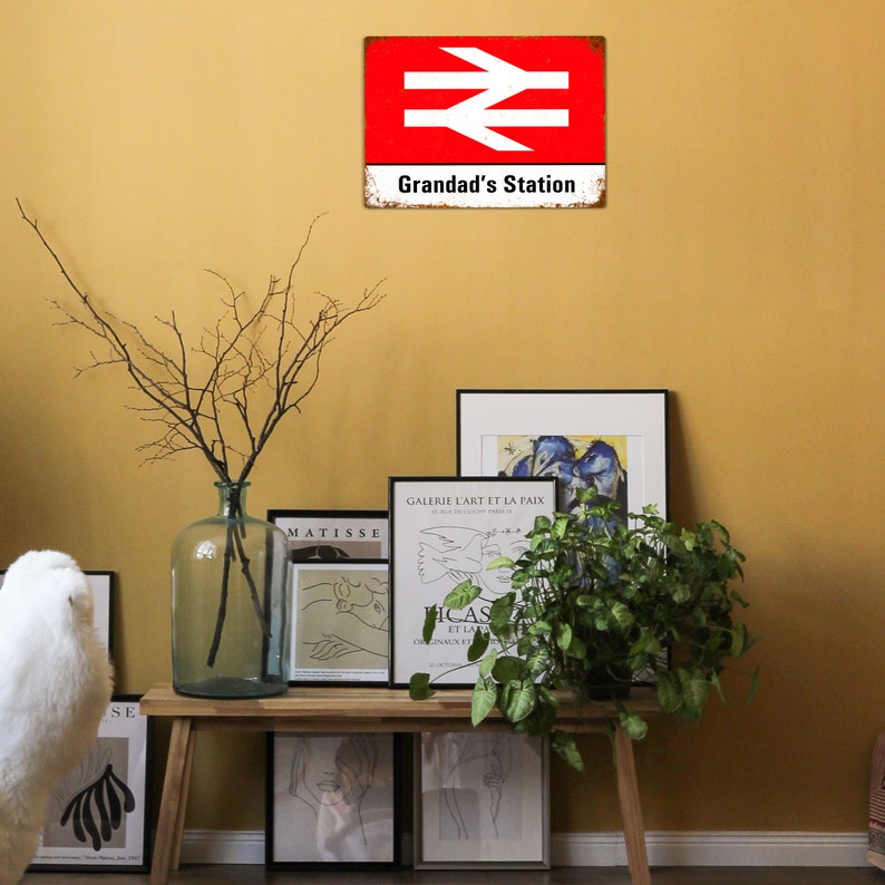 May include: A red and white vintage metal sign with the text "Grandad's Station" and a railway symbol. The sign is hanging on a yellow wall with a vase of branches and framed artwork on a wooden shelf below.