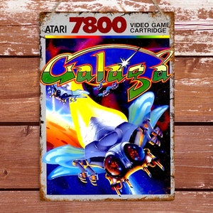 May include: A vintage Atari 7800 video game cartridge for the game Galaga. The cartridge is blue and white with a colorful image of a spaceship and a star. The text "VIDEO GAME CARTRIDGE" is printed on the cartridge.