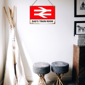 May include: A red and white metal sign with a railway symbol and the text "Dad's Train Room". The sign is hanging on a white wall with two gray woven stools in the foreground.