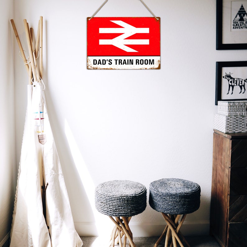 May include: A red and white metal sign with a railway symbol and the text "Dad's Train Room". The sign is hanging on a white wall with two gray woven stools in the foreground.