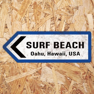 Puede incluir: Un letrero azul y blanco con una flecha negra apuntando hacia la izquierda. El letrero dice "Surf Beach, Oahu, Hawaii, USA".