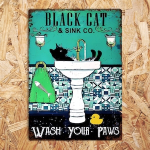K&ouml;nnte beinhalten: Ein Vintage-Schild aus Metall mit einer schwarzen Katze, die ihre Pfoten in einem wei&szlig;en Porzellanwaschbecken w&auml;scht. Das Schild lautet "Black Cat & Sink Co. Wash Your Paws".