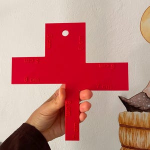 Puede incluir: Una plantilla de plástico de color rojo brillante en forma de cruz con varios recortes y bordes etiquetados con medidas en centímetros, incluyendo 5 cm, 6 cm, 7 cm y 8 cm, con un orificio circular en la parte superior vertical.