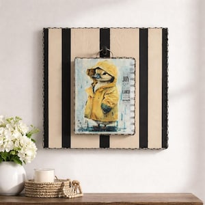 May include: A framed wall art piece featuring a duckling in a yellow raincoat standing in the rain. The artwork is mounted on a striped background with black and beige stripes. Text on the artwork reads "The Joy of the Lord is your strength."