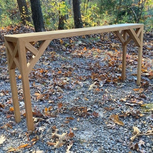 May include: A light-colored wooden console table with a rectangular top and decorative geometric supports. The table is approximately 60 inches long and stands on four legs. The design features an open, lattice-like pattern on the legs.