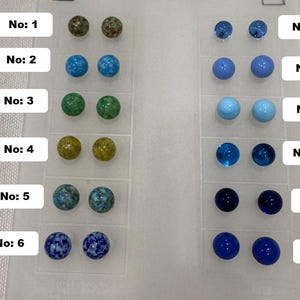 May include: A collection of twelve pairs of round stud earrings in various colors and patterns. The earrings are arranged in rows and columns, with each pair displayed in a clear plastic holder. The colors include green, blue, and yellow.