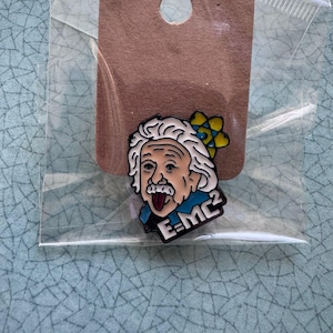 May include: A colorful enamel pin featuring a cartoon depiction of Albert Einstein with white hair, a mustache, and his tongue sticking out. The pin includes the formula E=MC² and a yellow and blue flower. The pin is attached to a brown card.