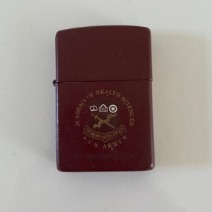 May include: A brown lighter with a gold emblem that reads 'Academy of Health Sciences, U.S. Army,  'Educating Fighting Strength''. The emblem also includes a house, a caduceus, and a star.