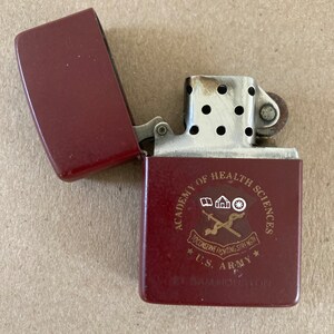 May include: A red lighter with a silver top and a gold inscription that reads "Academy of Health Sciences, To Conserve Fighting Strength, U.S. Army, Ft. Sam Houston".
