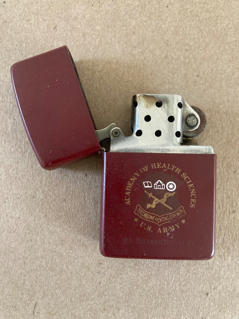 May include: A red lighter with a silver top and a gold inscription that reads "Academy of Health Sciences, To Conserve Fighting Strength, U.S. Army, Ft. Sam Houston".