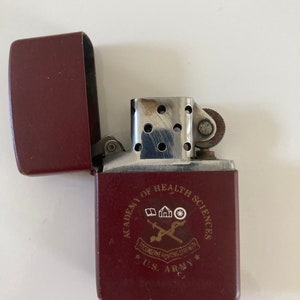 May include: A red lighter with a silver top and a gold inscription that reads "Academy of Health Sciences, U.S. Army,  Decisive Fighting Strength".