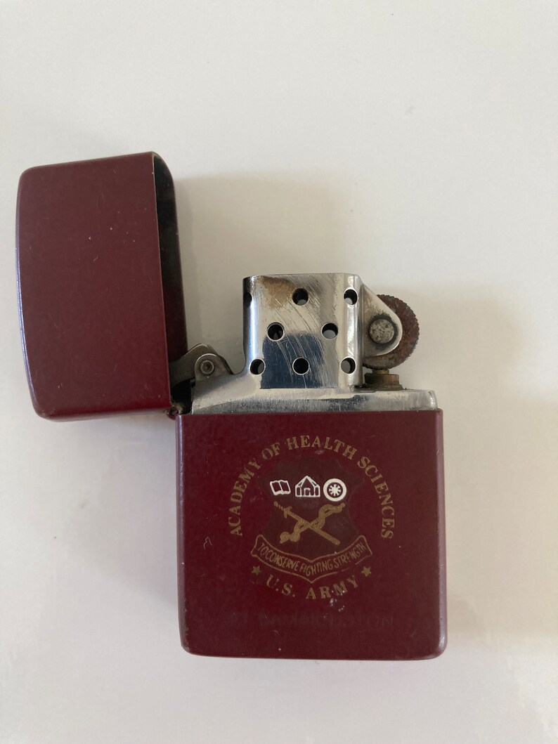 May include: A red lighter with a silver top and a gold inscription that reads "Academy of Health Sciences, U.S. Army,  Decisive Fighting Strength".