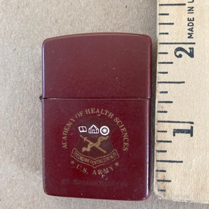 May include: A red lighter with a gold emblem that reads "Academy of Health Sciences To Conserve Fighting Strength U.S. Army Fort Sam Houston"
