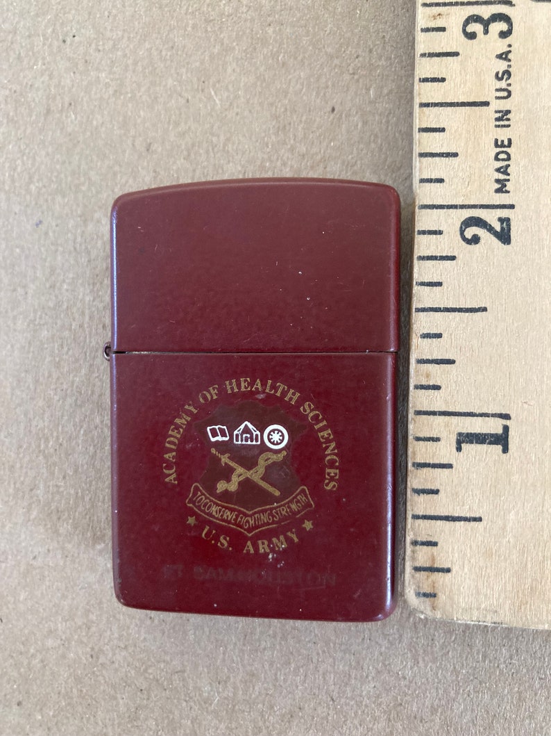 May include: A red lighter with a gold emblem that reads "Academy of Health Sciences To Conserve Fighting Strength U.S. Army Fort Sam Houston"