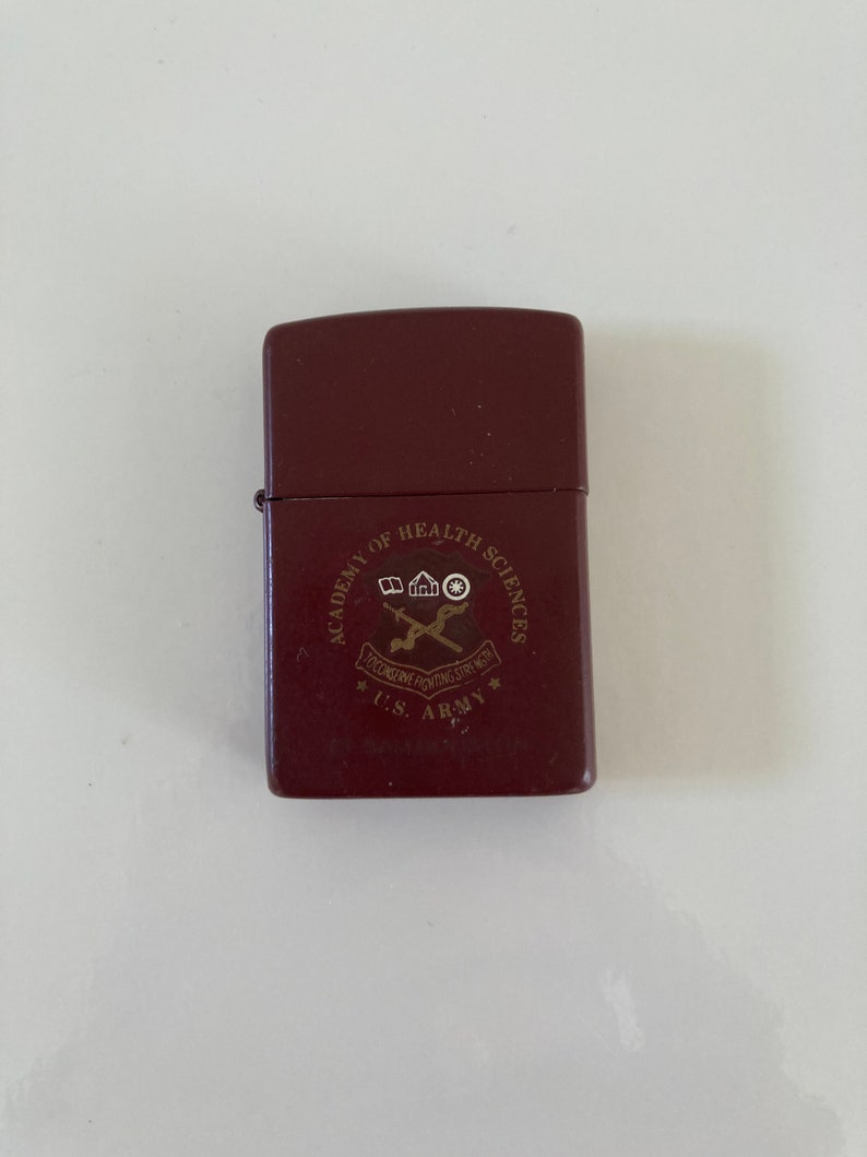 May include: A brown lighter with a gold emblem that reads 'Academy of Health Sciences, U.S. Army,  'Educating Fighting Strength''. The emblem also includes a house, a caduceus, and a star.