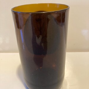 May include: A dark brown glass vase with a yellow rim. The vase is cylindrical in shape and has a smooth, glossy finish.