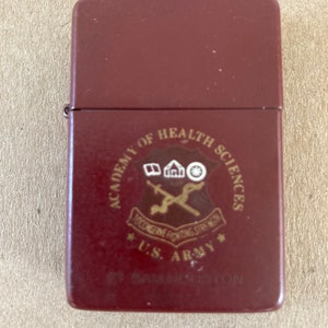 May include: A maroon lighter with a gold emblem of the U.S. Army Academy of Health Sciences. The emblem features a caduceus, a building, and a wheel.