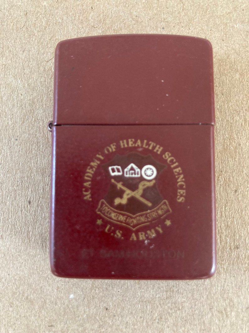 May include: A maroon lighter with a gold emblem of the U.S. Army Academy of Health Sciences. The emblem features a caduceus, a building, and a wheel.