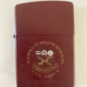 May include: A maroon lighter with a gold emblem that reads "Academy of Health Sciences,  *U.S. Army*" with a crossed sword and a building with a book, a house, and a wheel.