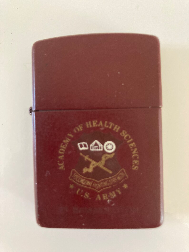 May include: A maroon lighter with a gold emblem that reads "Academy of Health Sciences,  *U.S. Army*" with a crossed sword and a building with a book, a house, and a wheel.