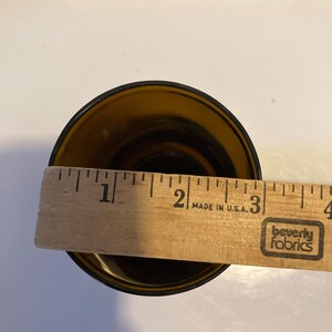 May include: A brown glass drinking glass with a diameter of approximately 2 inches. The glass is sitting on a white surface with a wooden ruler behind it. The ruler has the words "Made in U.S.A." and "Beverly Fabrics" printed on it.