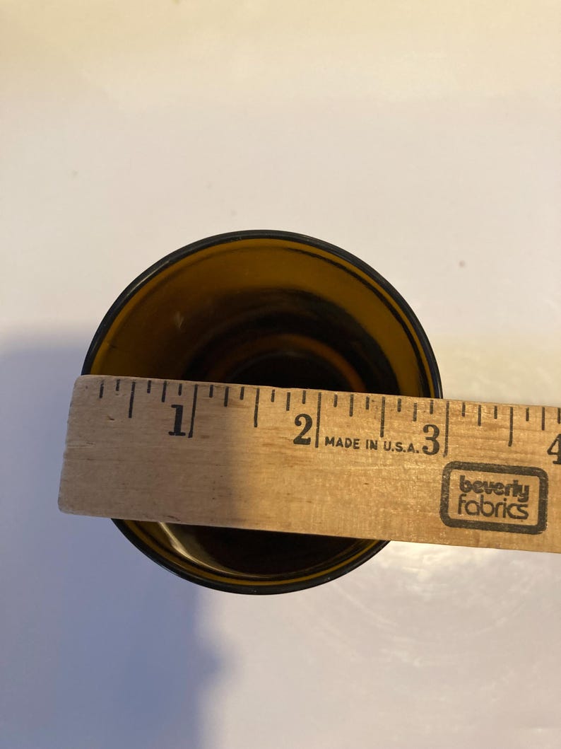 May include: A brown glass drinking glass with a diameter of approximately 2 inches. The glass is sitting on a white surface with a wooden ruler behind it. The ruler has the words "Made in U.S.A." and "Beverly Fabrics" printed on it.