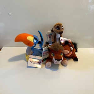 May include: Four Disney plush toys: a toucan with an orange beak, a meerkat, a lion cub, and a warthog. Each plush has a Disney Store tag attached. The toys are in various shades of brown, orange, blue, and yellow.