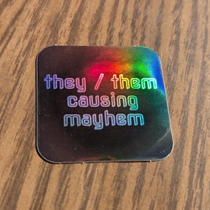 May include: A square sticker with rounded corners, featuring the text "they / them causing mayhem" in a rainbow gradient font. The background has a holographic, iridescent effect, with colours shifting across the surface.