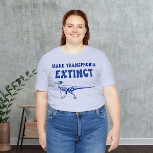 May include: A light gray t-shirt with the text "Make Transphobia Extinct" in blue lettering. A blue skeletal dinosaur is printed below the text.