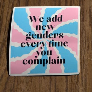 May include: A sticker featuring a blue, pink, and white design with the text "We add new genders every time you complain" in black. The background has a radial pattern. The sticker is rectangular.