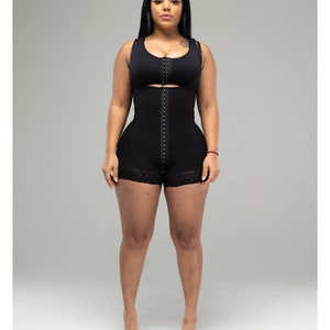 Peut inclure: Gainant noir avec soutien-gorge intégré et short. Le vêtement est doté d'une fermeture à agrafes sur le devant et d'une bordure en dentelle sur le short. Le modèle se tient devant un fond gris. La marque "SNATCHED" est visible en bas.
