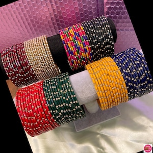 May include: A selection of colourful bangles, including red, gold, black, and blue, each decorated with small, sparkling embellishments. The bangles are displayed on black and white stands, showcasing their vibrant hues and intricate details.
