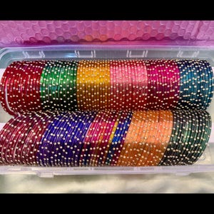 Könnte beinhalten: Eine Sammlung bunter Armreifen in einer transparenten Kunststoffbox. Die Armreifen sind in verschiedenen Farben erhältlich, darunter Rot, Grün, Gelb, Rosa, Blau und Orange, und jeweils mit kleinen, weißen, gepunkteten Verzierungen versehen. Die Armreifen sind in Reihen gestapelt.