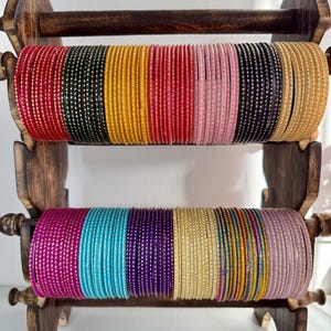 May include: A two-tiered wooden display stand holding rows of thin metal bangles in various colours including red, green, yellow, pink, black, beige, magenta, turquoise, purple, and lavender, each featuring a repeating pattern of small gold dots.