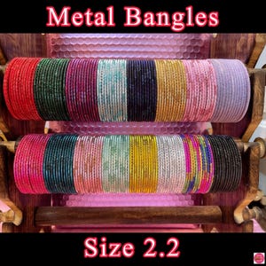 Op de afbeelding: Een set van 24 metalen armbanden in verschillende kleuren, waaronder roze, groen, goud, zilver en paars. De armbanden zijn in twee rijen op een houten standaard gerangschikt. De tekst "Metal Bangles" en "Size 2.2" is zichtbaar in de afbeelding.