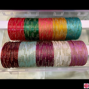 May include: Rows of colorful glass bangles in shades of red, green, yellow, and teal, and purple, white, and pink. The bangles are displayed in a clear plastic container, showcasing their vibrant colors and delicate design.