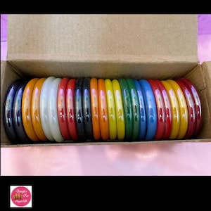 May include: A box of 18 colourful glass bangles in a rainbow of colours, including red, orange, yellow, green, blue, and purple. The bangles are arranged in a row in the box, with the colours alternating.