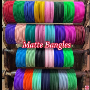 May include: A collection of colorful matte bangles stacked on a wooden stand. The bangles are arranged in rows of different colors, including red, green, blue, yellow, pink, and purple. The text "Matte Bangles" is displayed in the center of the image.