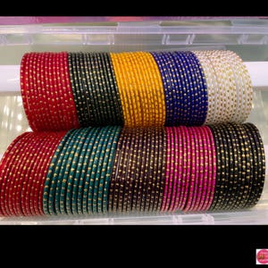 May include: A selection of vibrant glass bangles in a range of colours, such as red, black, yellow, blue, and white. Each bangle is decorated with tiny, gold-coloured dots. The bangles are neatly arranged, displaying a variety of colours and designs.