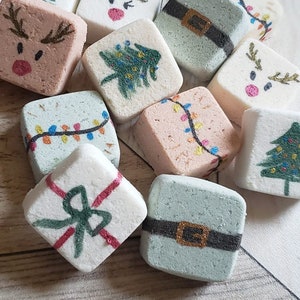 May include: A collection of square, pastel-colored bath bombs with Christmas-themed designs. The designs include a Christmas tree, reindeer faces, gift boxes, and strings of lights. Red ornaments and a pinecone are in the background.