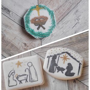 May include: A collection of Christmas-themed decorative stones. One stone features a gold cross and a manger scene within a green wreath. Two other stones depict nativity scenes with gold star accents, in black and white.