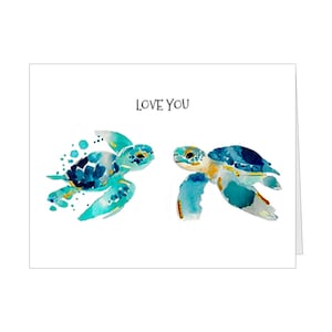 May include: A white greeting card featuring a watercolor illustration of two sea turtles swimming toward each other in shades of teal, blue, and yellow. The text LOVE YOU is printed in black, centered above the two sea turtles on the card.