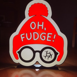 May include: A novelty sign featuring a red knit cap with a white outline and the words "OH, FUDGE!" in white. Below the cap are black-rimmed glasses with one cracked lens. The sign is illuminated.