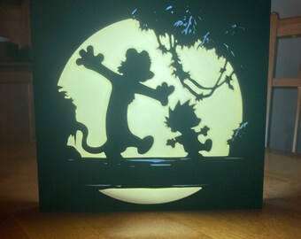 May include: A black wooden silhouette of Garfield and Odie standing on a branch in front of a full moon. The silhouette is lit from behind, creating a glowing effect.