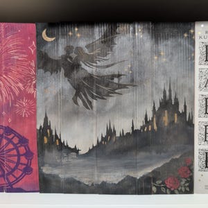 May include: A bookshelf with three books and a decorative panel. The panel features a dark illustration of a winged figure over a castle, with a crescent moon and stars. The book on the right is titled "BABEL".