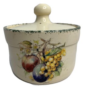 May include: A cream-colored ceramic sugar bowl with a matching lid featuring a knob handle. The exterior displays a colorful botanical illustration of plums, grapes, and white blossoms, finished with a dark green speckled rim along the top edge.