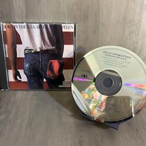 May include: A compact disc and case for Bruce Springsteen's "Born in the U.S.A." album. The CD is silver with black text, and the case features an image of a person in jeans and a white shirt with a red and white striped background.