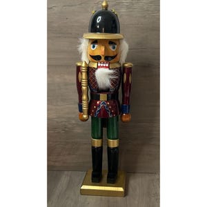 May include: A wooden nutcracker figure wearing a black helmet with gold trim, a red jacket with gold and blue accents, and green trousers. The figure features a white faux fur beard, painted facial details, and stands on a gold base.