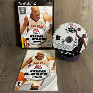 以下が含まれることがあります： PlayStation 2のビデオゲーム「NBA Live 2004」で、表紙にはバスケットボール選手が描かれています。ゲームケース、ディスク、インサートが見えます。ゲームのロゴとプレイヤーの画像が目立ちます。