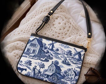 May include: A rectangular shoulder bag featuring a blue and white toile de jouy pattern depicting rural scenes with houses, windmills, and figures. The bag has a black leather strap with gold-tone hardware, a top zipper closure, and black trim.